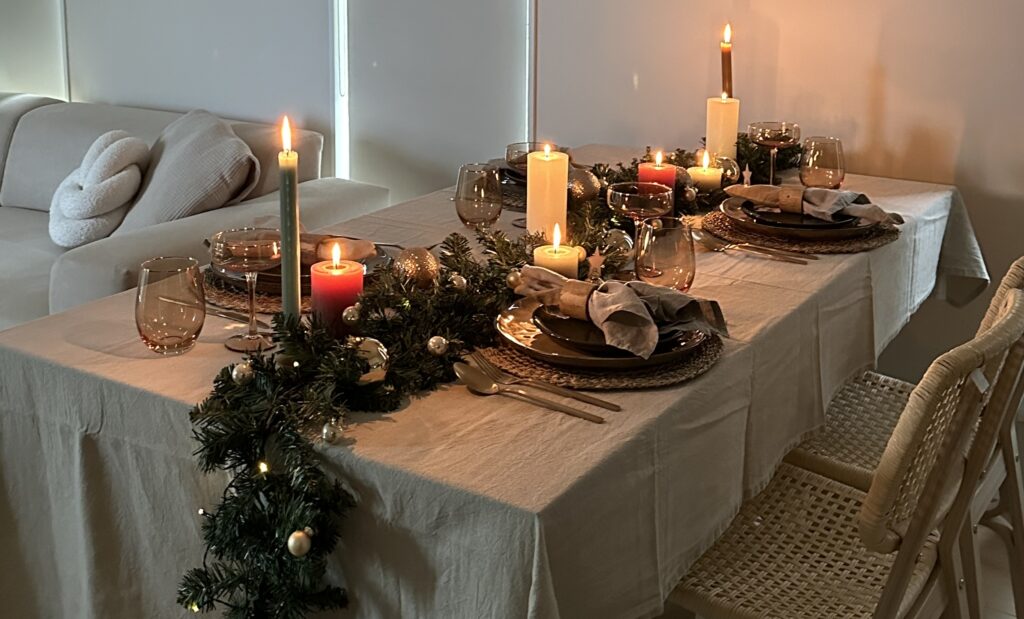 Een gedekte kersttafel met beige tafelkleed, slinger met lichtjes, gouden bestek en kaarsen